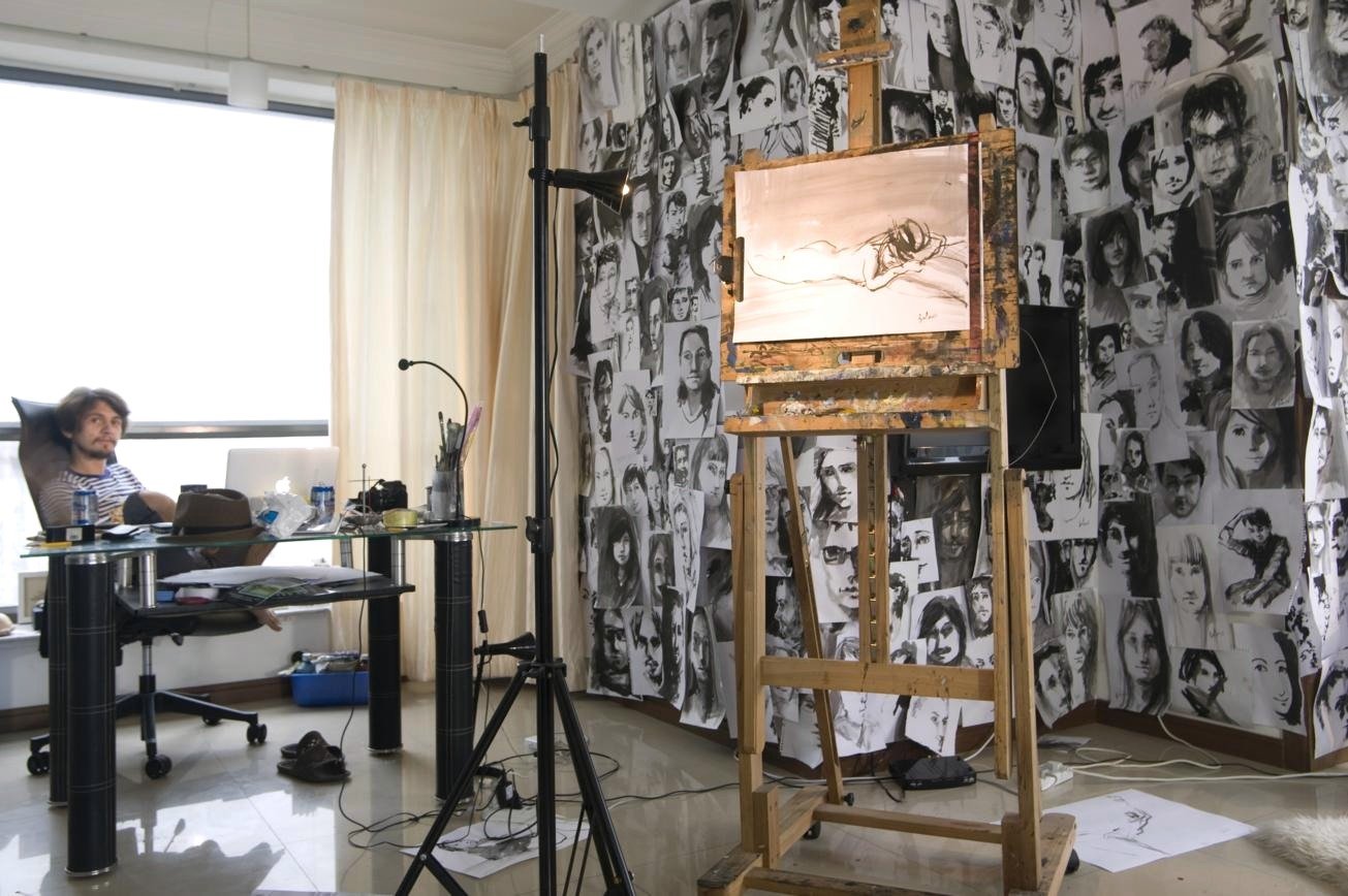 An artist at a desk beside an easel and a wall covered with painted portraits