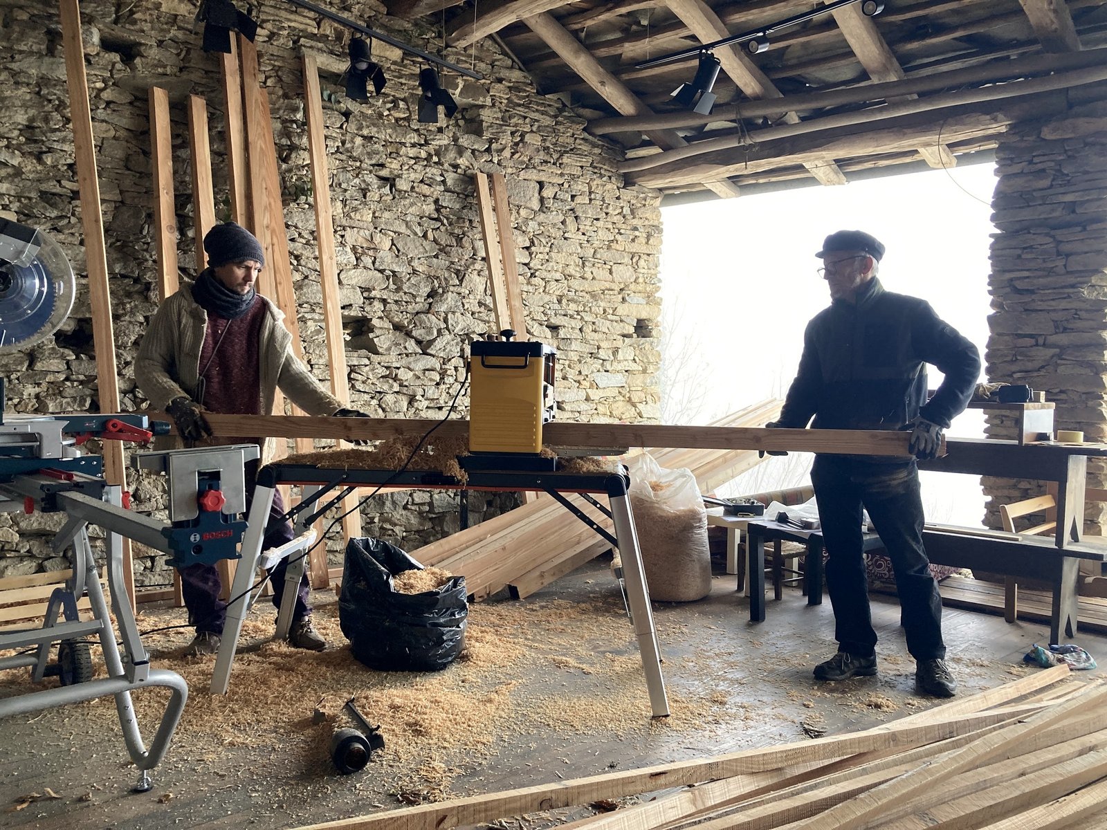 Two people working a timber beam in a stone-walled workshop